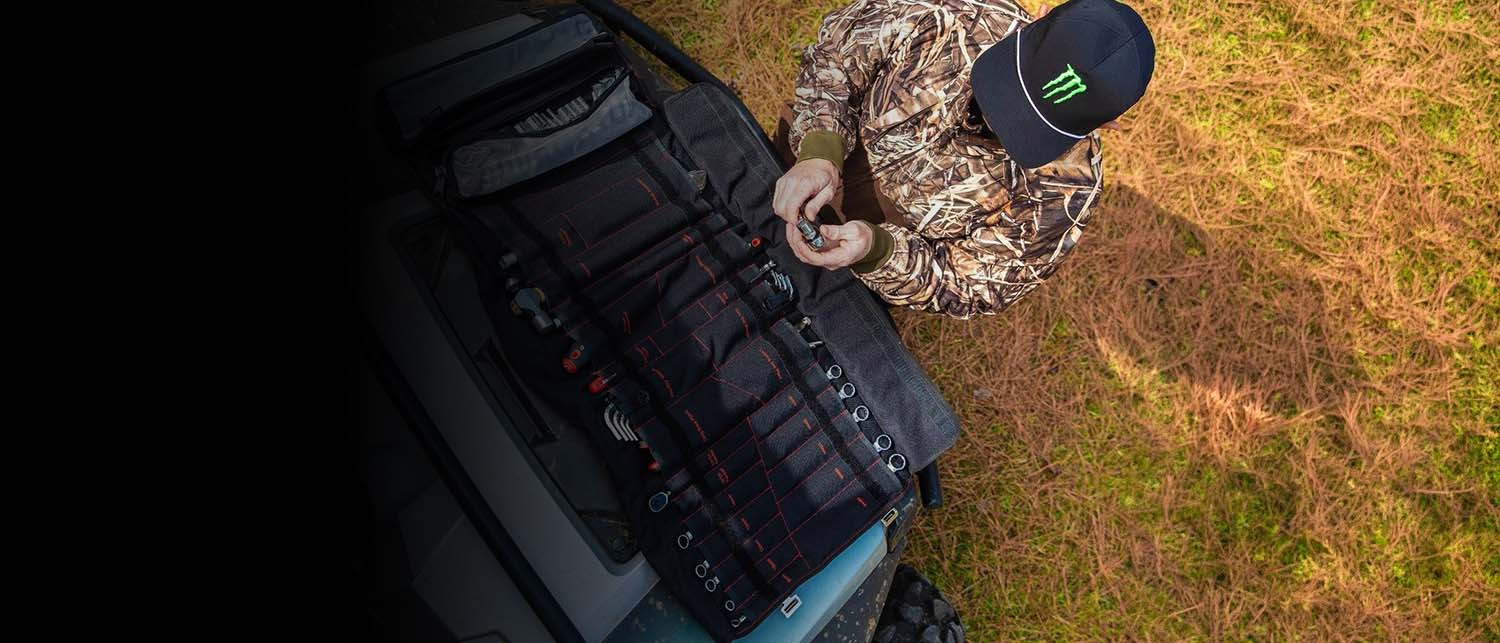 A person in camouflage clothing and a black cap with a green Monster Energy logo stands beside a vehicle trunk containing a large black tackle organizer bag filled with fishing gear, outdoors on grass.