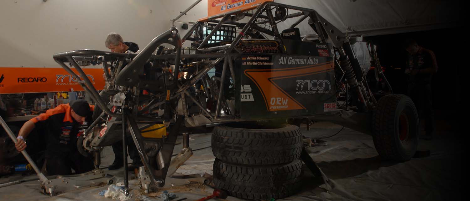 AGM Class 1 race buggy with exposed suspension and stacked tires in a garage, with crew members working around it. The vehicle has prominent sponsor decals including Micron, DEW, and 3D German Auto.