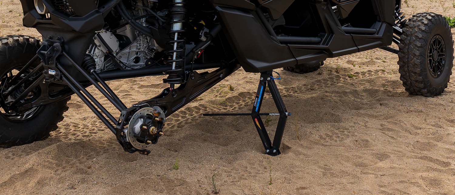 A black Can Am Maverick X3 supported by an AGM Scissor Jack on sandy ground.