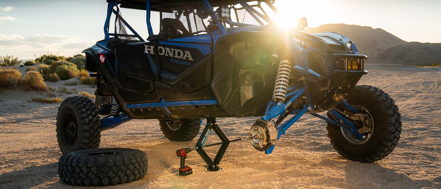 A blue and black Honda Talon off-road vehicle raised on an AGM Scissor jack in a desert setting at sunset, with a spare tire on the ground and a cordless tool nearby.