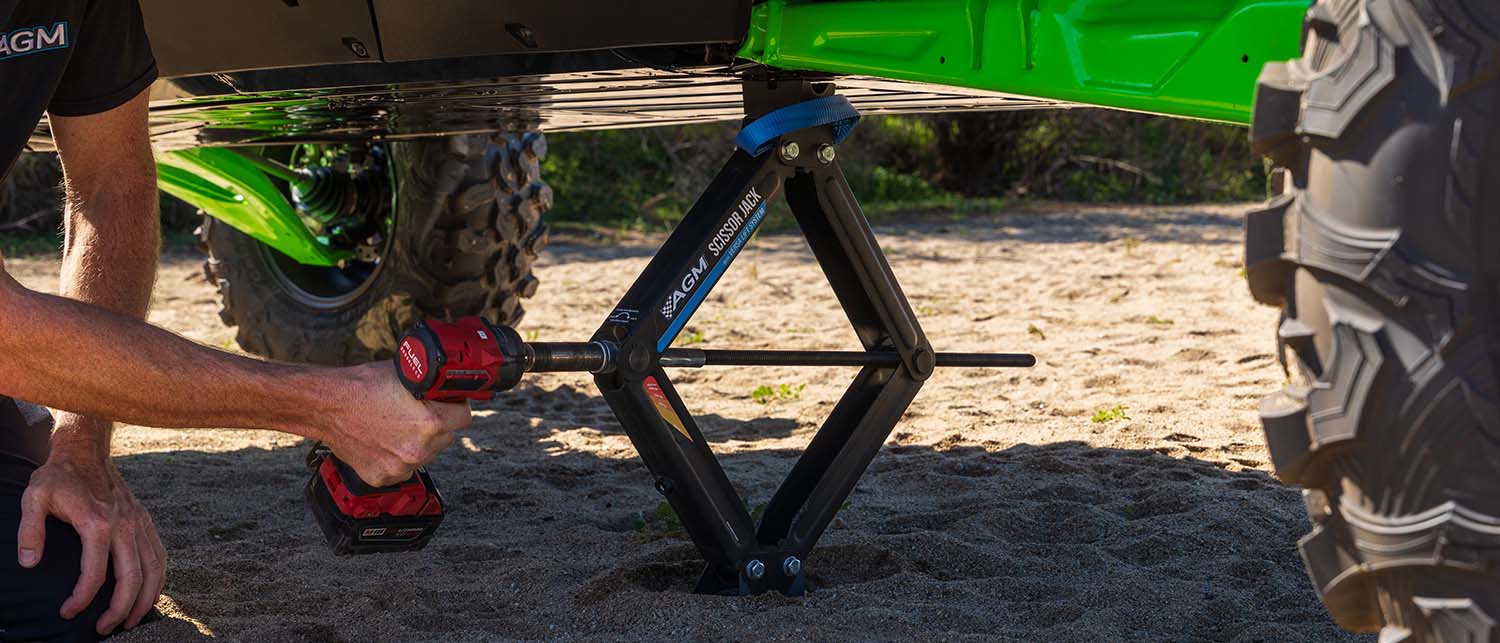 A man using a red cordless impact driver to raise an AGM Scissor Jack that is supporting a green and black Kawasaki H2 SXS on sand.