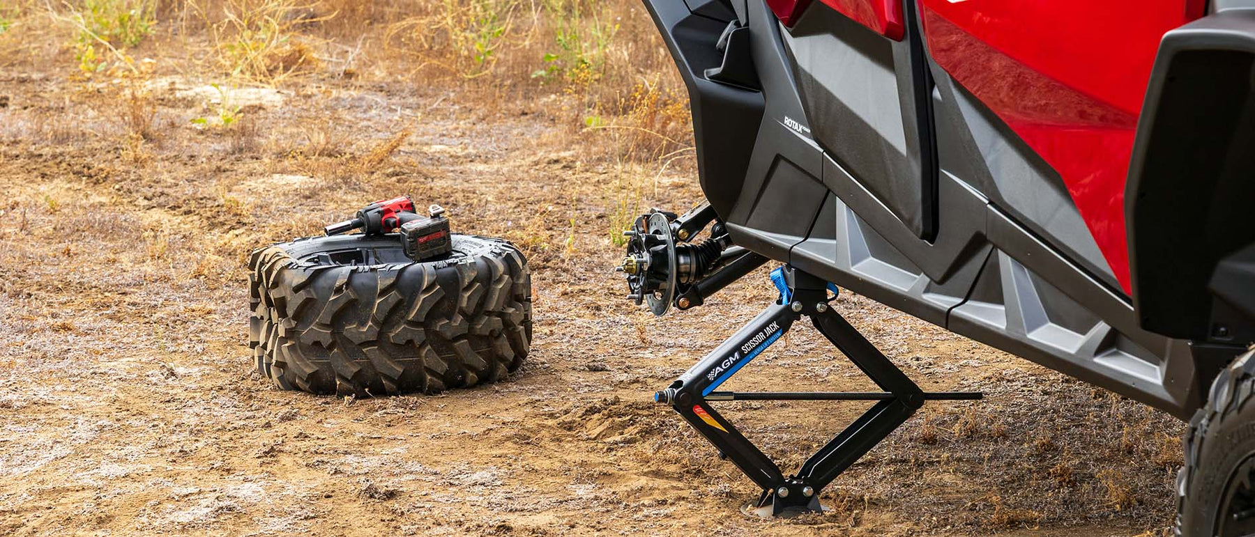 A red and black off-road vehicle is lifted by an AGM scissor jack on a dirt ground, with a spare tire lying nearby on the ground.