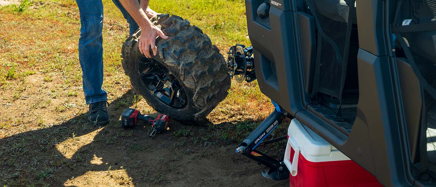 A person outdoors removes a large off-road tire from a black Can Am Defender using an AGM Scissor jack and impact wrench. The vehicle is parked on grass and dirt ground next to a red and white cooler.