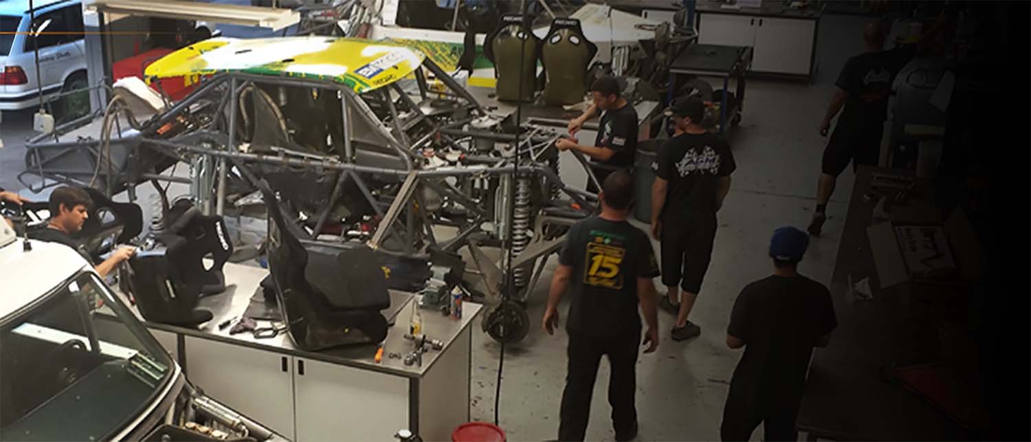 A group of people working inside a garage or workshop around a stripped metal race car frame, with tools and equipment visible on workbenches.