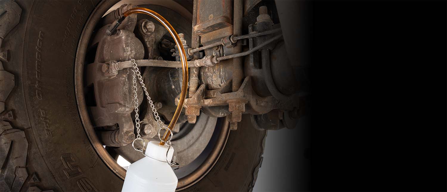 A close-up of a vehicle wheel hub and brake assembly with a clear plastic bottle connected by a chain and orange hose, likely for tire change or fluid transfer, set against a dark background.