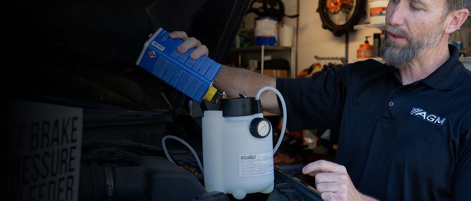 A man in a dark navy polo shirt with an AGM logo is pouring liquid from a blue container into an AGM Brake Pressure Bleeder bottle connected to a trucks brake system inside a garage.
