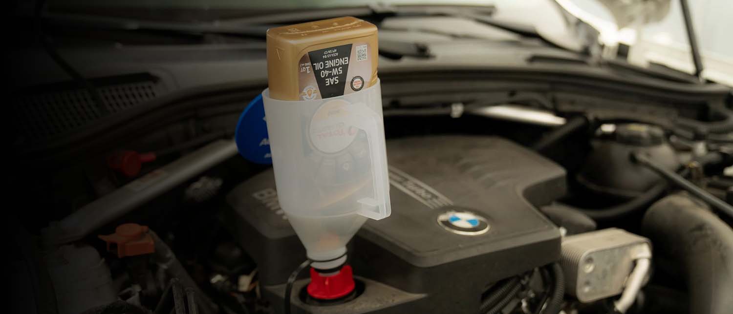 A bottle of BMW engine oil with a clear plastic funnel and red cap placed in an open car engine bay.