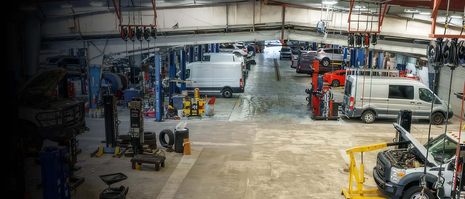 A wide interior view of an auto repair shop with multiple service bays, vehicles, tools, lifts, and equipment under industrial ceiling fixtures.
