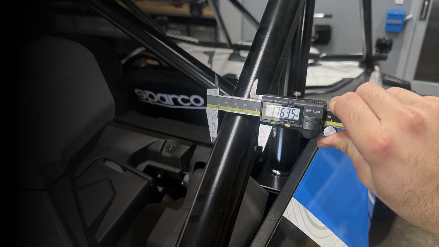 A hand using a digital caliper to measure the width of an offroad vehicle roll bar inside a race car cockpit.