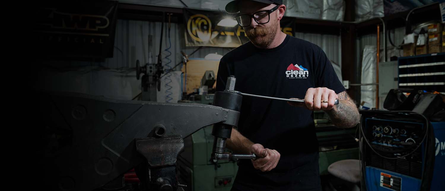 A man wearing a black t-shirt with a 'Climb The Mountain' logo works in a workshop environment, using AGM Uni ball tool on a lower control arm