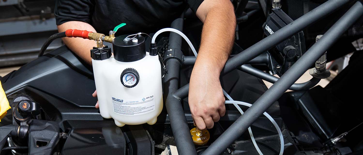A person using an AGM Brake Pressure Bleeder with hoses connected to a black engine or machinery component in a real-world setting.