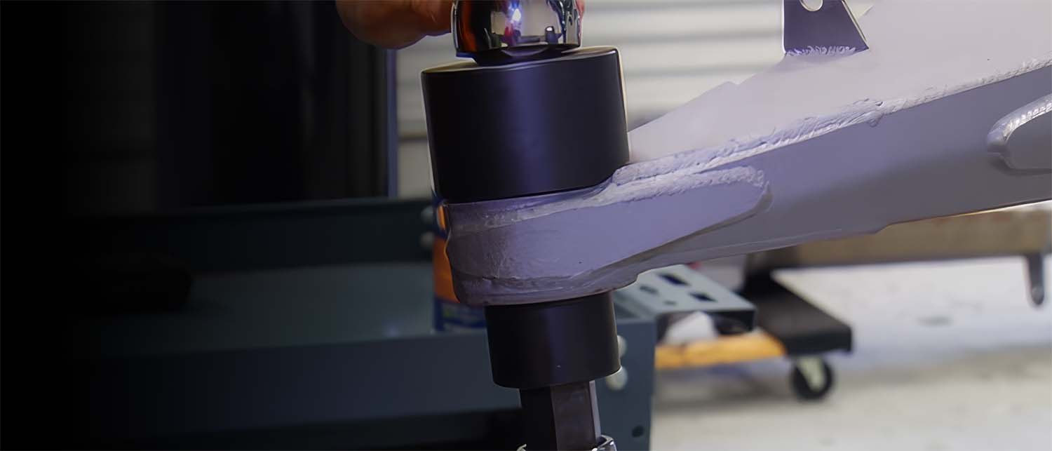 A close-up shot of an AGM Uni-Ball tool on a gray metal lower control arm, with a hand partially visible at the top in an race workshop setting.