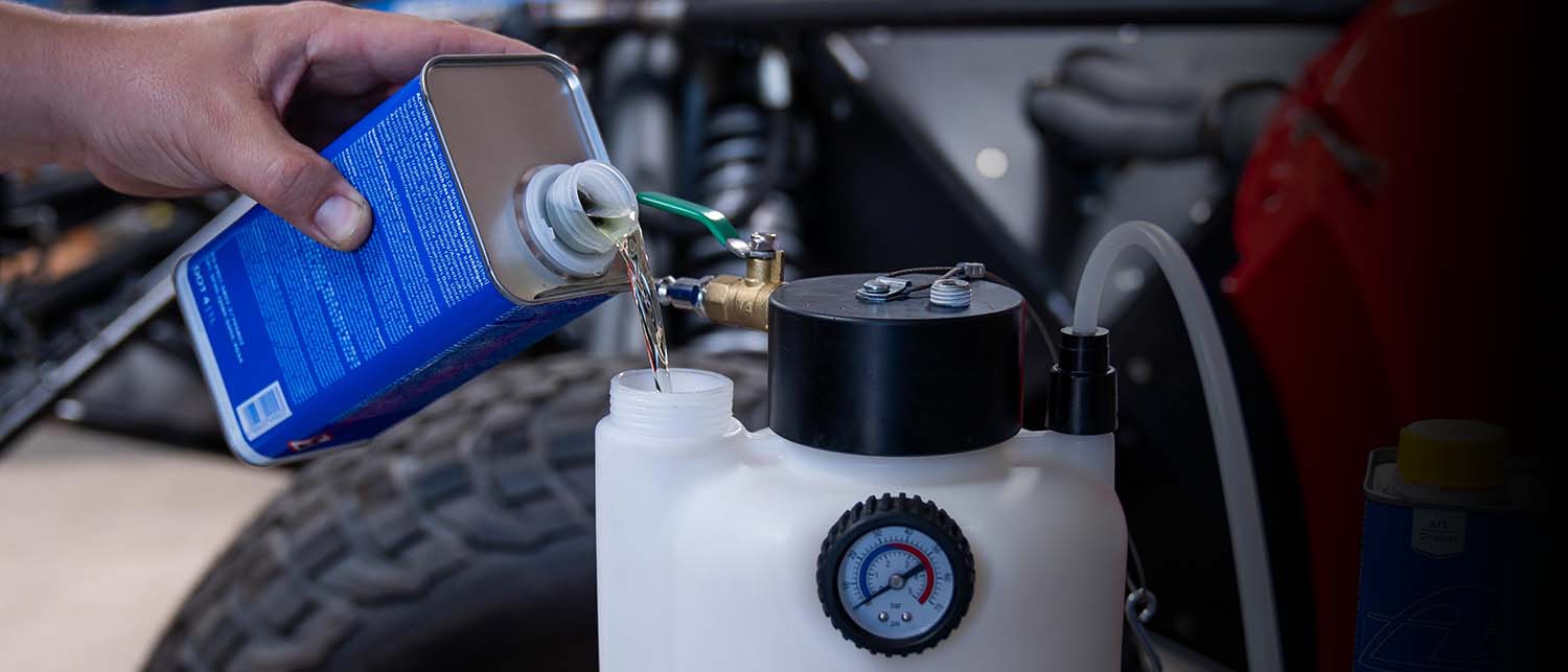 A hand pouring liquid from a blue metal can into a white pressure tank connected to a hose, with a motorcycle engine and tire in the background.