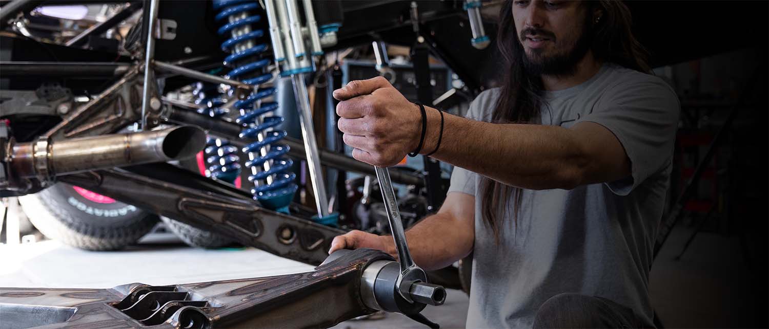 A man in a gray t-shirt works on a vehicle suspension using the AGM Uni-Ball tool in a garage setting.