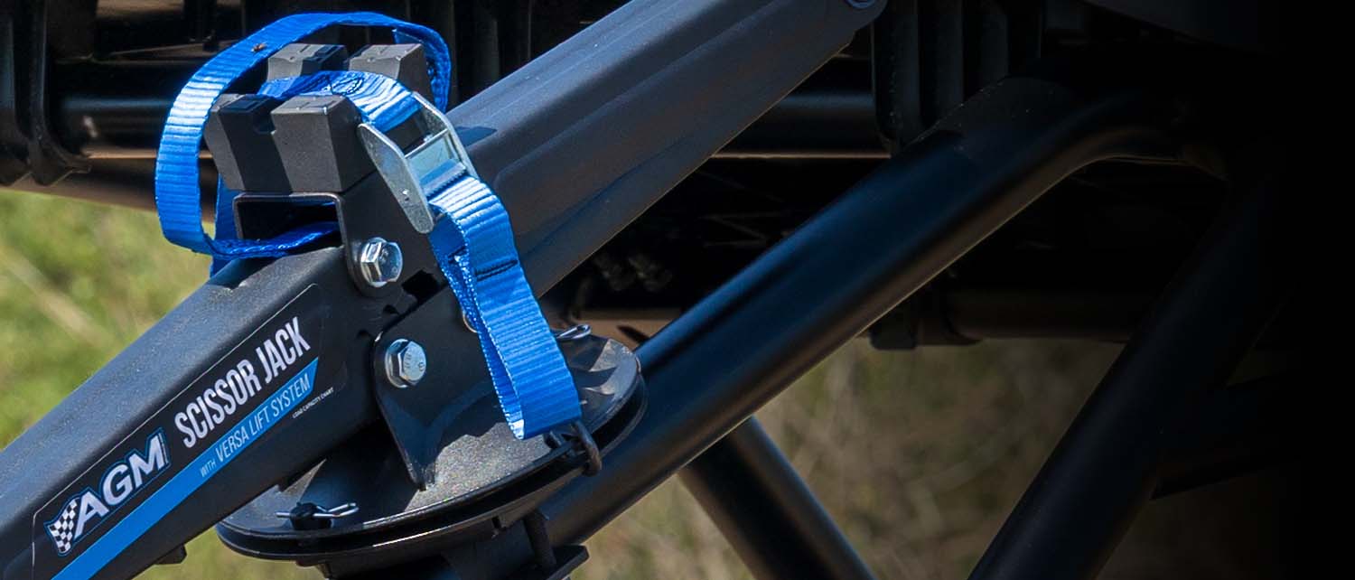 Close-up of an AGM scissor jack strapped into an AGM Scissor jack Mount attached to a black vehicle frame, set against a dark background with a hint of outdoor grass.