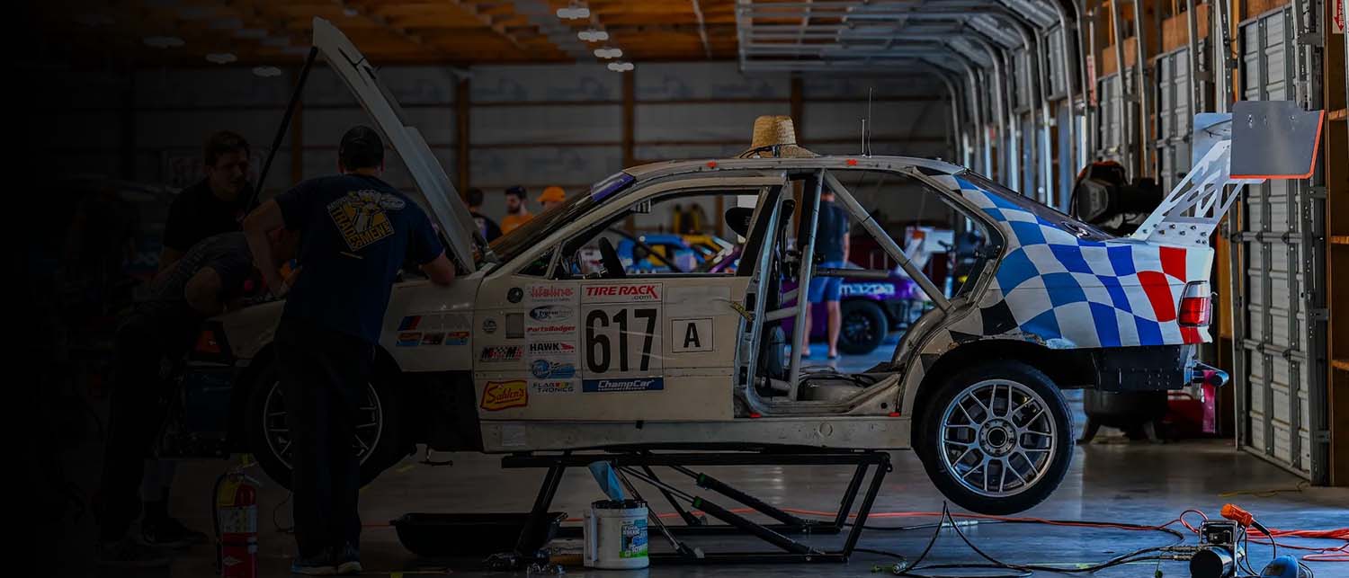 A white race car with blue and red checkered graphics and number 617 sits inside a garage or workshop, with people and equipment visible around it.